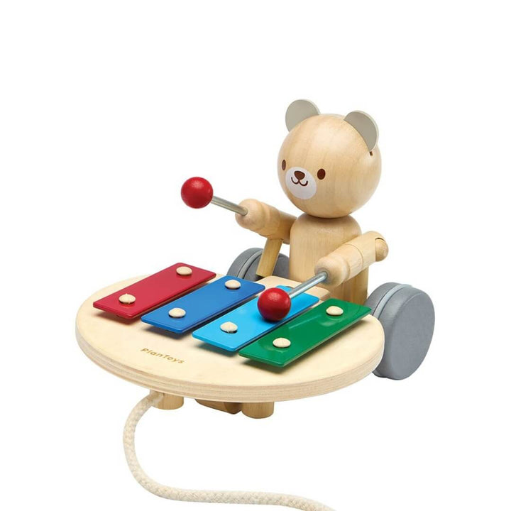 Wooden PlanToys pull-along bear tapping a colorful xylophone with red mallets, mounted on wheels with a pull cord attached. Bella Luna Toys.