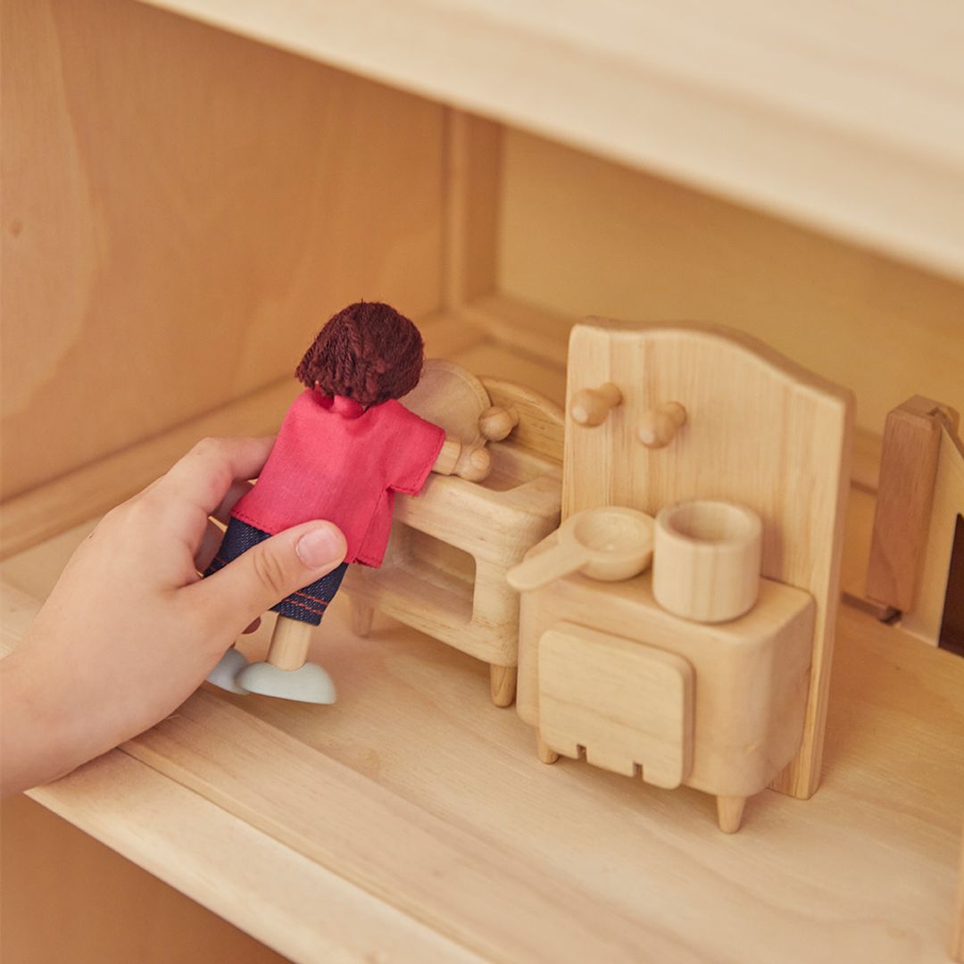Close-up of hands placing tiny wooden kitchen furniture in a dollhouse, showcasing detailed craftsmanship. The hand is holding a dollhouse doll with a pink shirt on and jeans and black hair. 