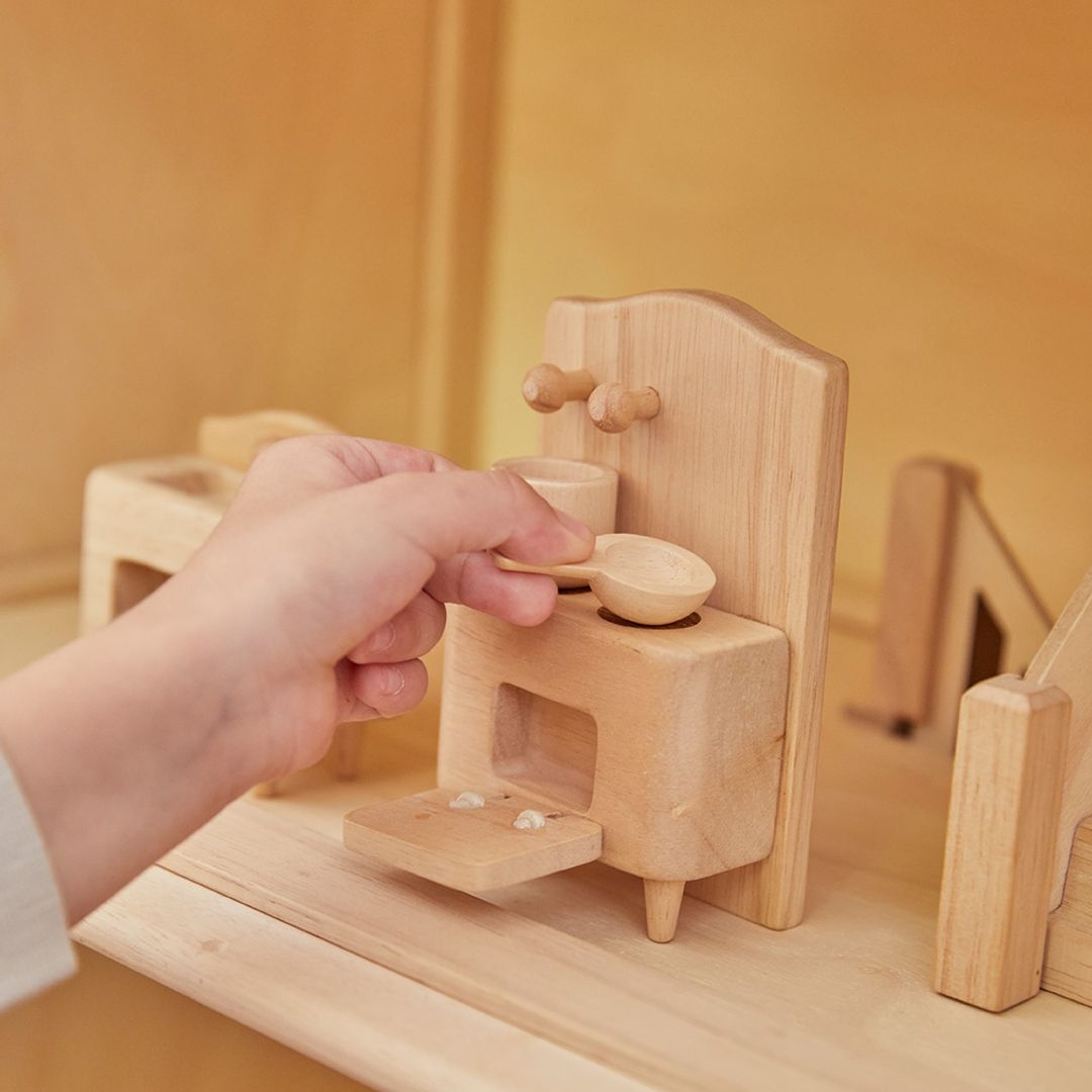 Close-up of hands placing a tiny wooden frying pan on a wooden stove amidst other kitchen furniture in a dollhouse, showcasing detailed craftsmanship.