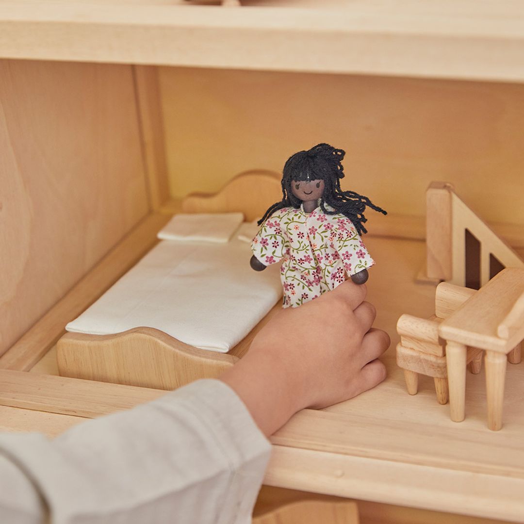 Close-up of hand placing a dollhouse doll with dark skin and black hair in a bedroom and near other wooden furniture in a dollhouse, showcasing detailed craftsmanship