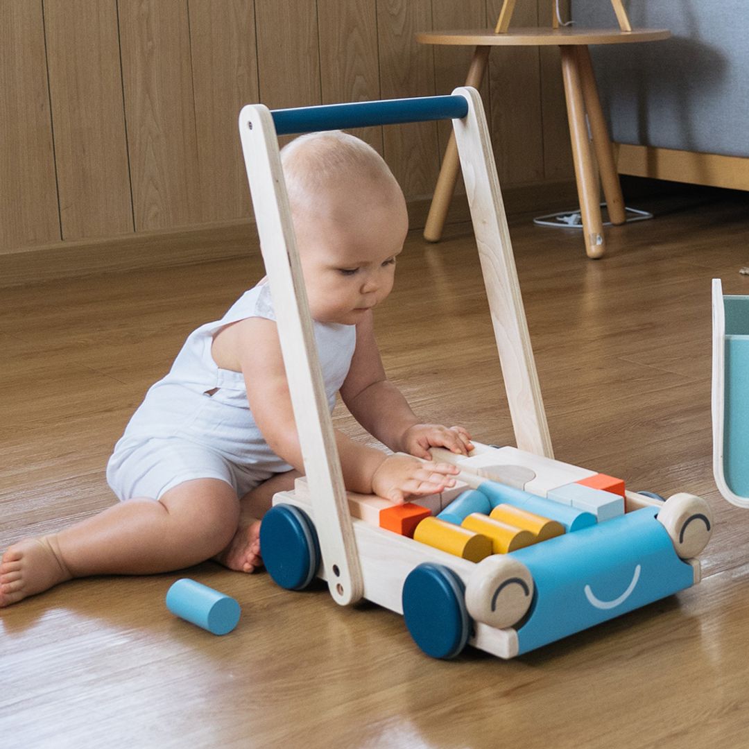 A one year old  in overalls sitting next to the walker wagon to pick up colorful wooden blocks from a PlanToys Baby Walker, a wooden push toy designed to support early walking.