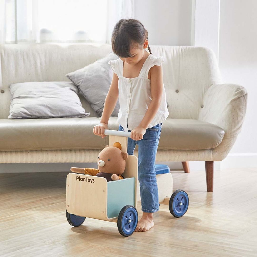 A child standing over the seat of the PlanToys Delivery Bike, a wooden ride-on toy with the front storage container filled with a teddy bear as she grips the handlebars.