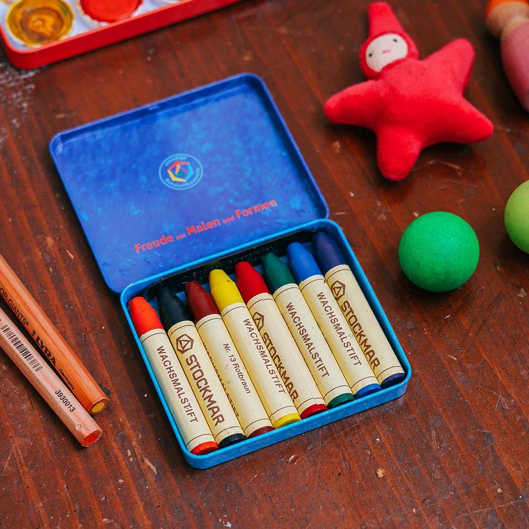 Set of Stockmar crayons in a blue box on a wooden surface with toys around