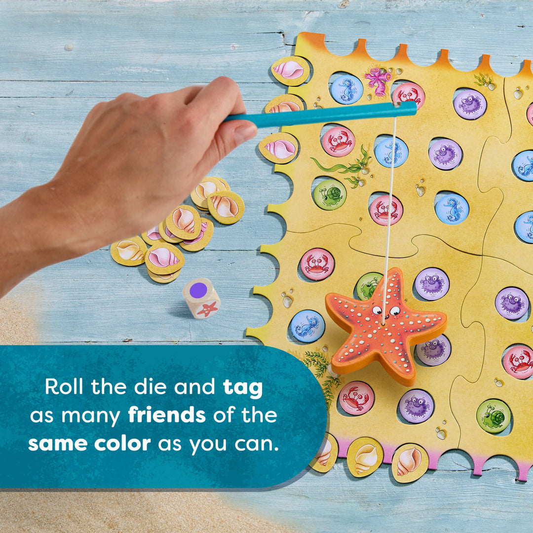 Colorful game board with pieces and dice, hand holding a stick with a dangling starfish, on a wooden surface.