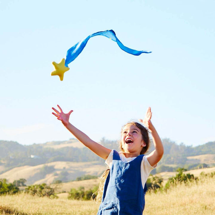 Child in a blue dress reaching towards a star-shaped skytail in a field with mountains in the background