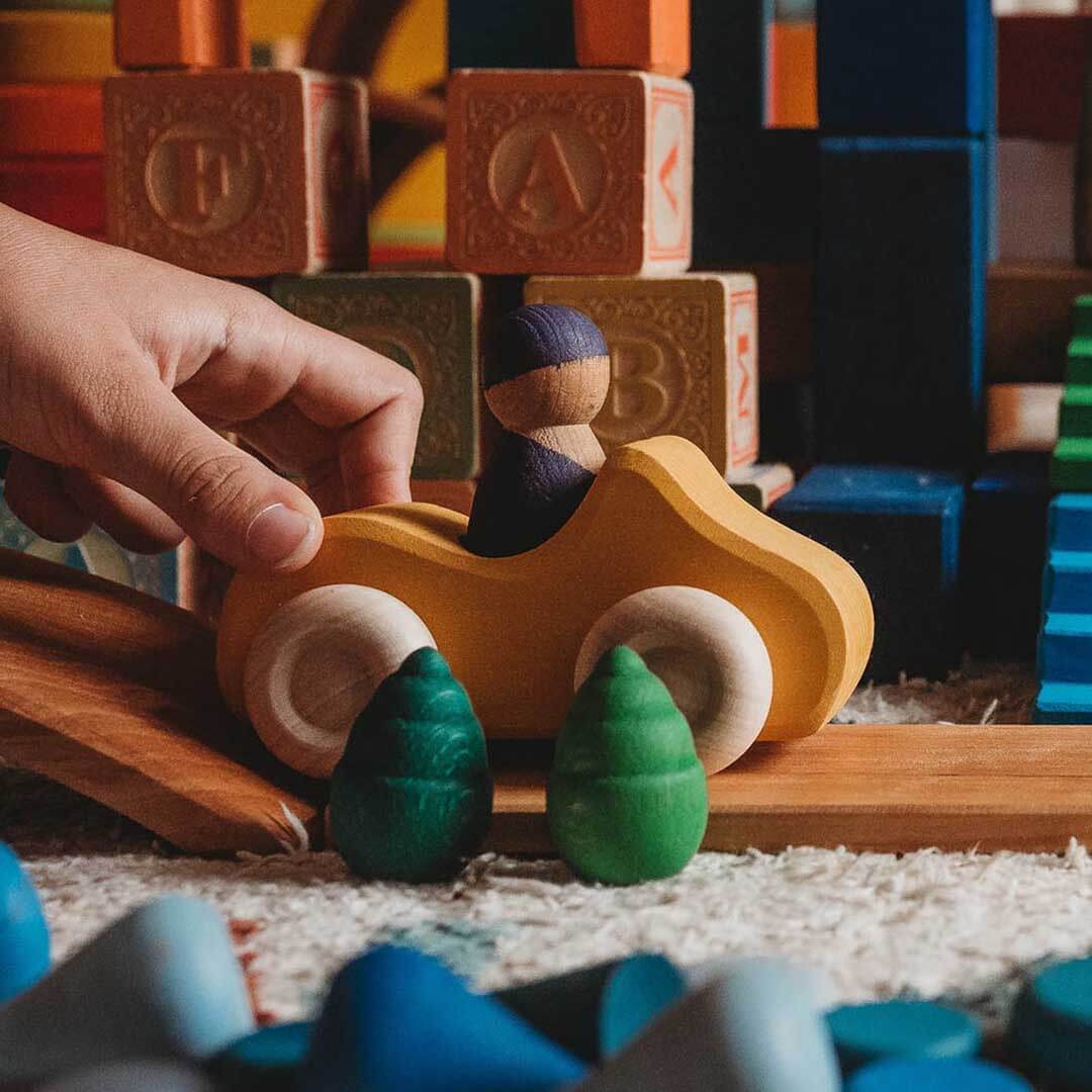 Hand interacting with a wooden toy car among other toys on a carpeted floor.