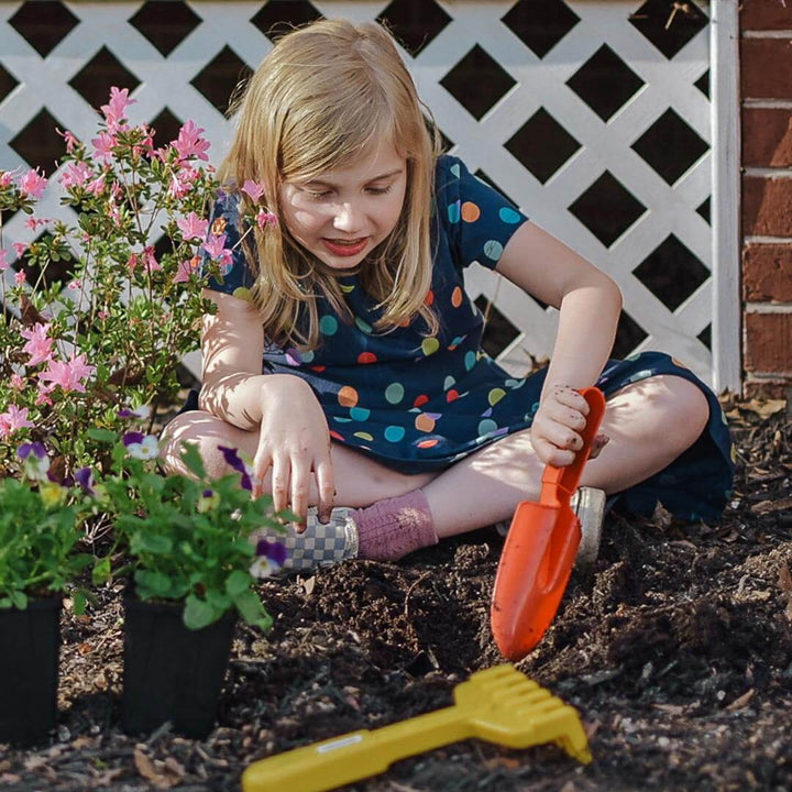 Child digging in mulch with red Spielstabil spade and yellow hand rake with pink flowers in the background