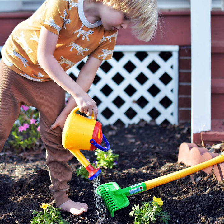 Boy pouring water from yellow watering can with long handled garden rake laying on the ground in a garden