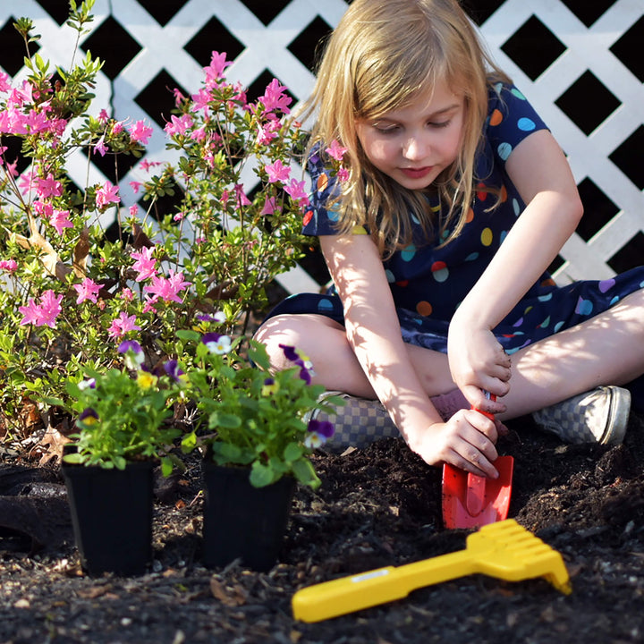 Girl digging in the garden with red spade and yellow hand rake laying on the ground