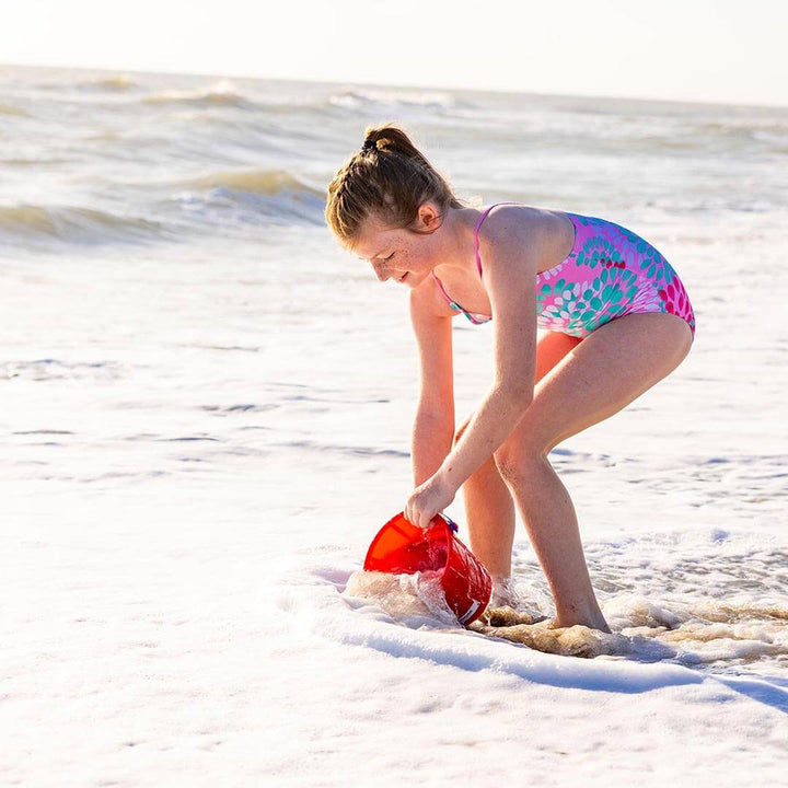 Girl standing in the ocean scooping water with Spielstabil Sand Pail in red