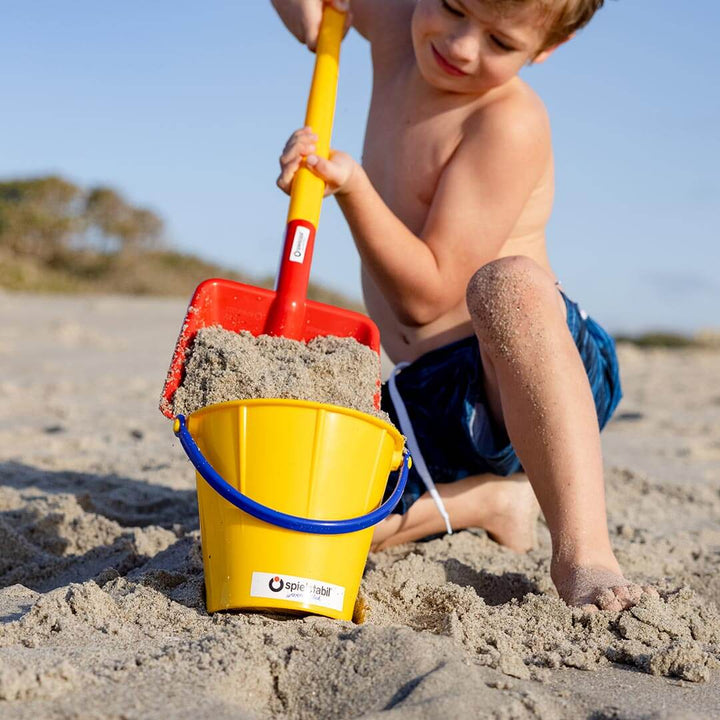 Boy kneeling on the beach shoveling sand into a yellow Spielstabil Sand Pail