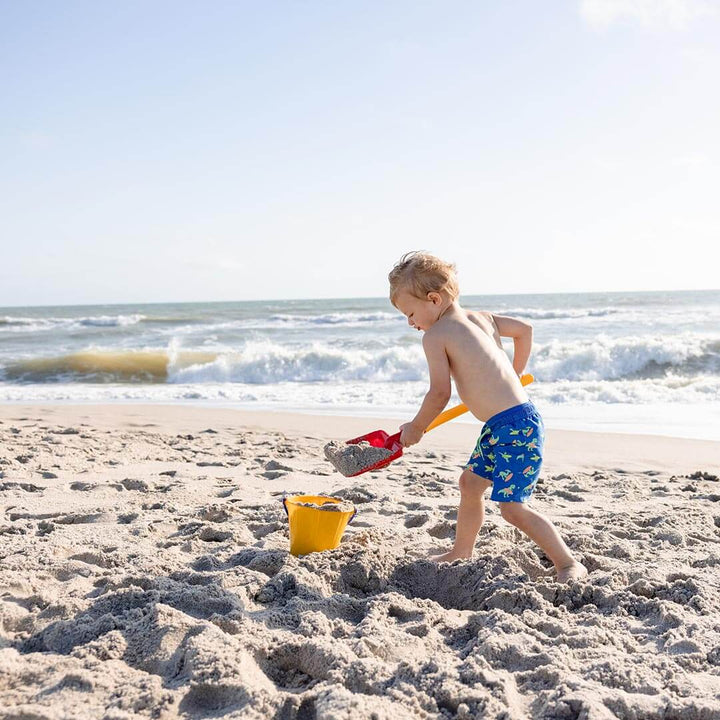 Boy standing in the sand by the ocean shoveling sand into a yellow Spielstabil Pail