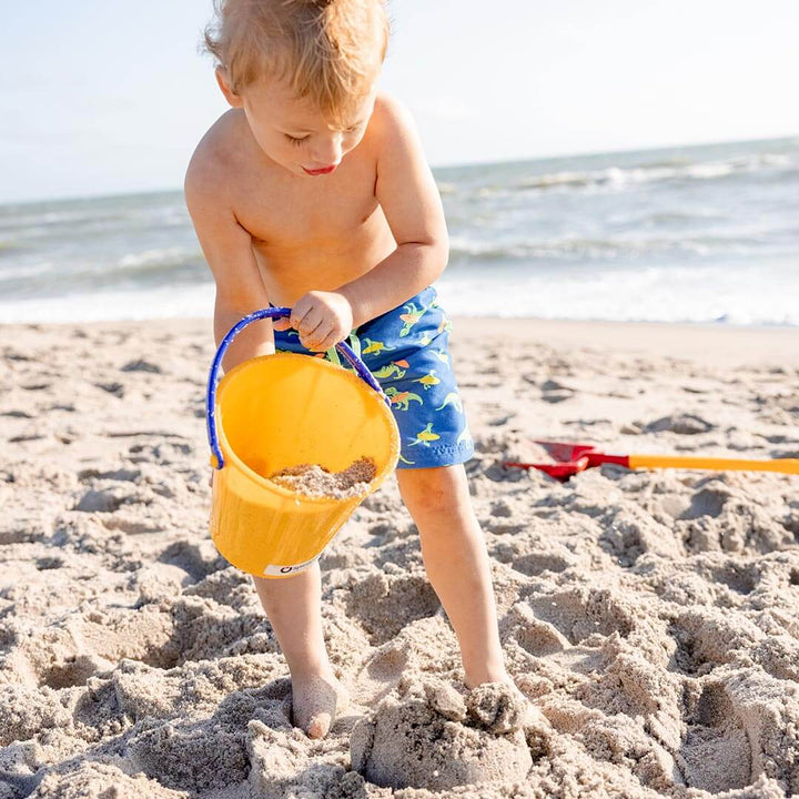 Boy standing in the sand at the ocean dumping sand from a yellow Spielstabil Pail