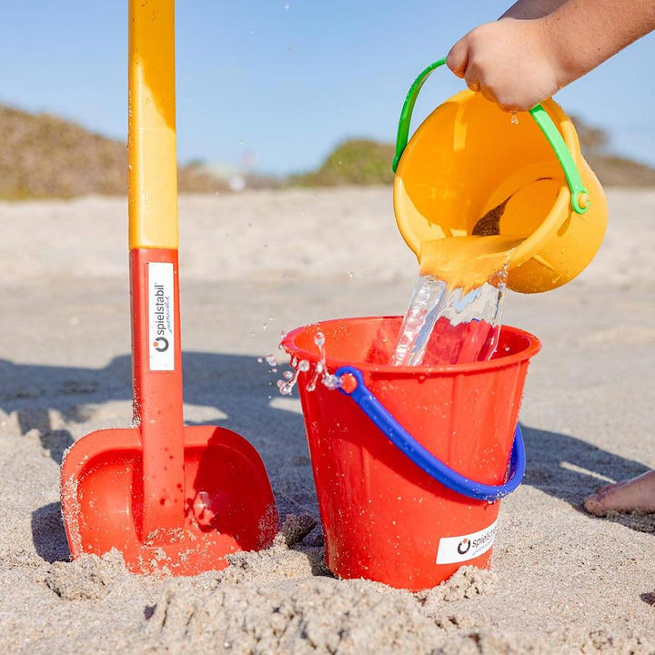 Spielstabil Long Handled Heavy Duty Beach Shovel in the sand with child pouring water from yellow pail into a red pail
