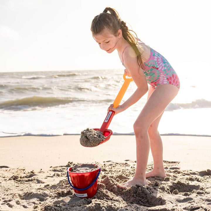 Child standing in front of the ocean shovel sand with a Spielstabil Long Handled Heavy Duty Beach Shovel into a red pail