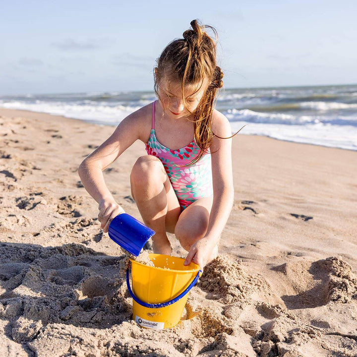 Girl kneeling in front of the ocean scooping sand into yellow Spielstabil Pail