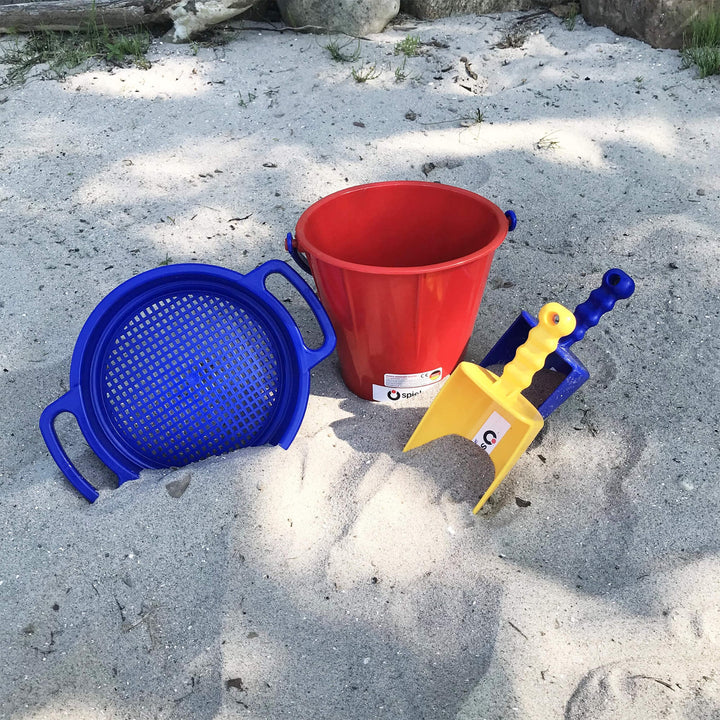 Spielstabil 4 Piece Large Sand Toys Bundle sitting in sand with blue sieve, red pail, yellow scoop, and blue scoop