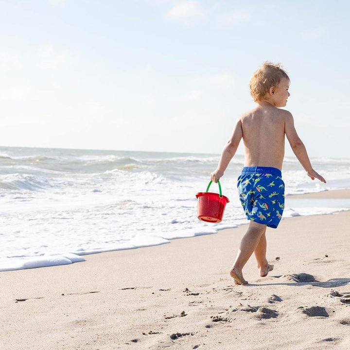 Young boy holding a red Spielstabil Small Sand Pail walking on the beach