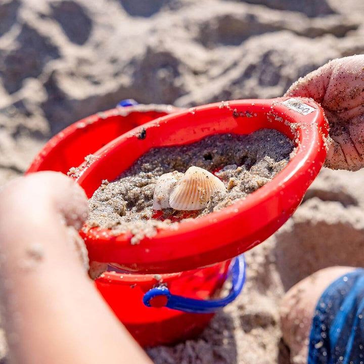 Child holding Spielstabil Small Sand Sieve in red filled with sand and shells at the beach