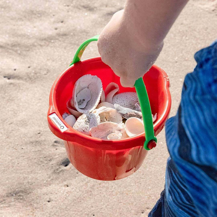 Child holding a red Spielstabil pail filled with beach shells