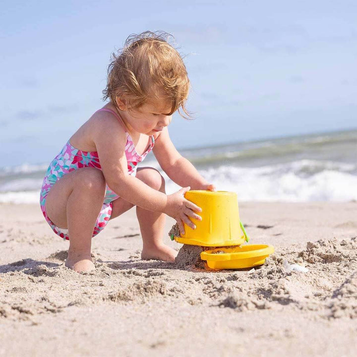 A child bending over an upside down yellow pail with yellow sand sieve on the beach