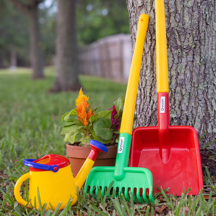 Spielstabil Watering Can sitting in the grass in front of garden rake and shovel leaning against a tree in the yard