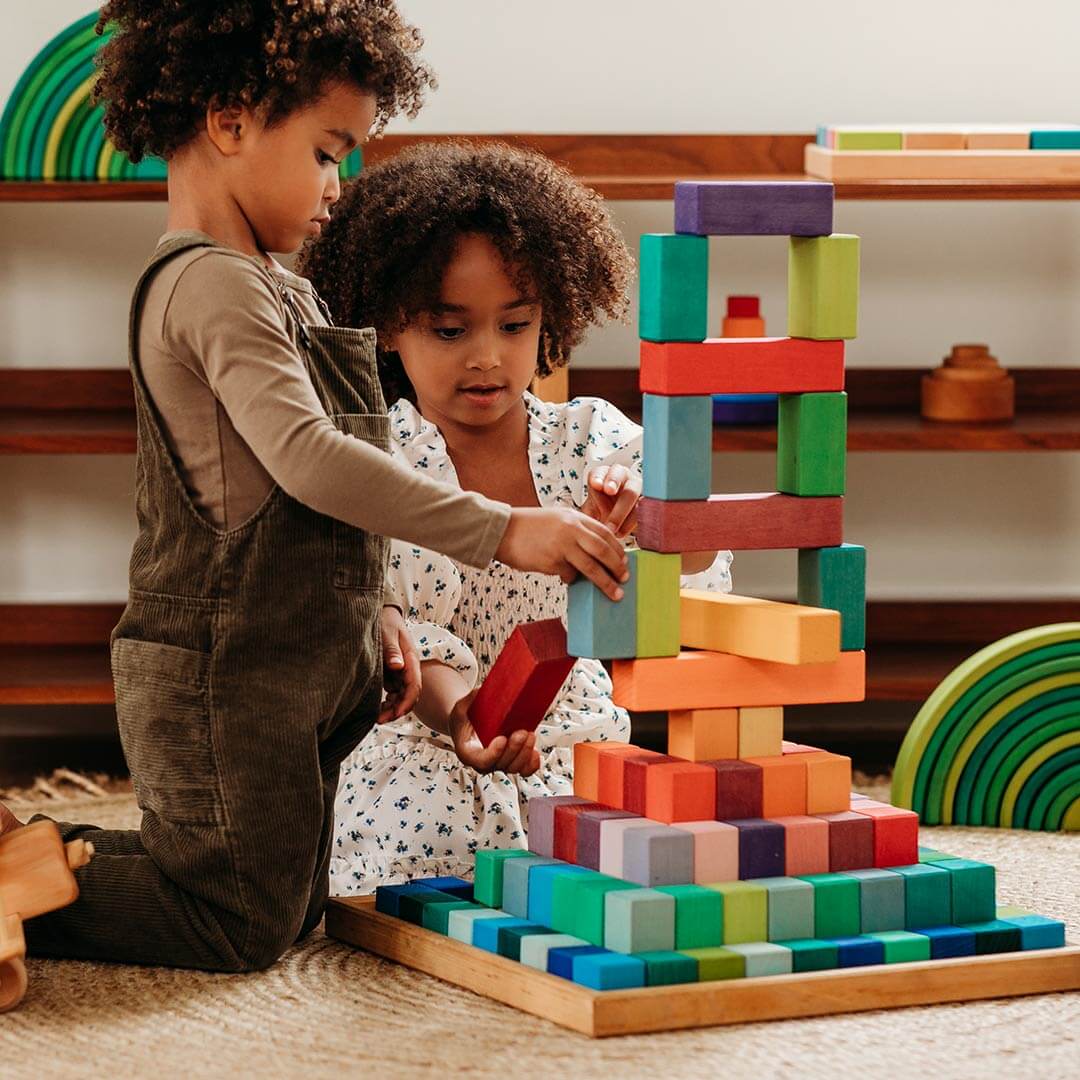 Two kids are playing with rainbow wooden building blocks in a colorful playroom.