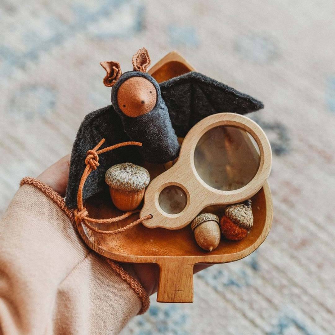 A hand holds a small wooden acorn-shaped tray holding a dark felt bat, acorns, and a magnifying glass, against a soft blurred background.