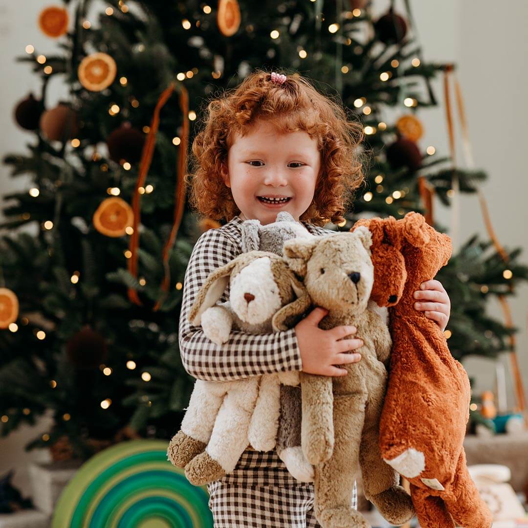 A young girl with curly red hair smiles while holding three plush stuffed animals in front of a decorated Christmas tree. The children's clothing is a black-and-white gingham pattern, and the background shows a warm, festive holiday scene.