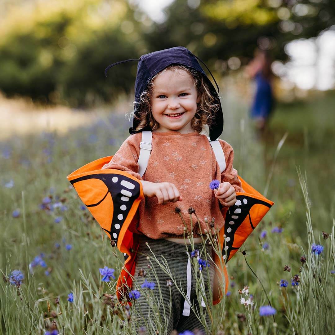 A young child smiling in a grassy field of purple wildflowers, wearing an orange and black butterfly costume with a dark hood and white shoulder straps.