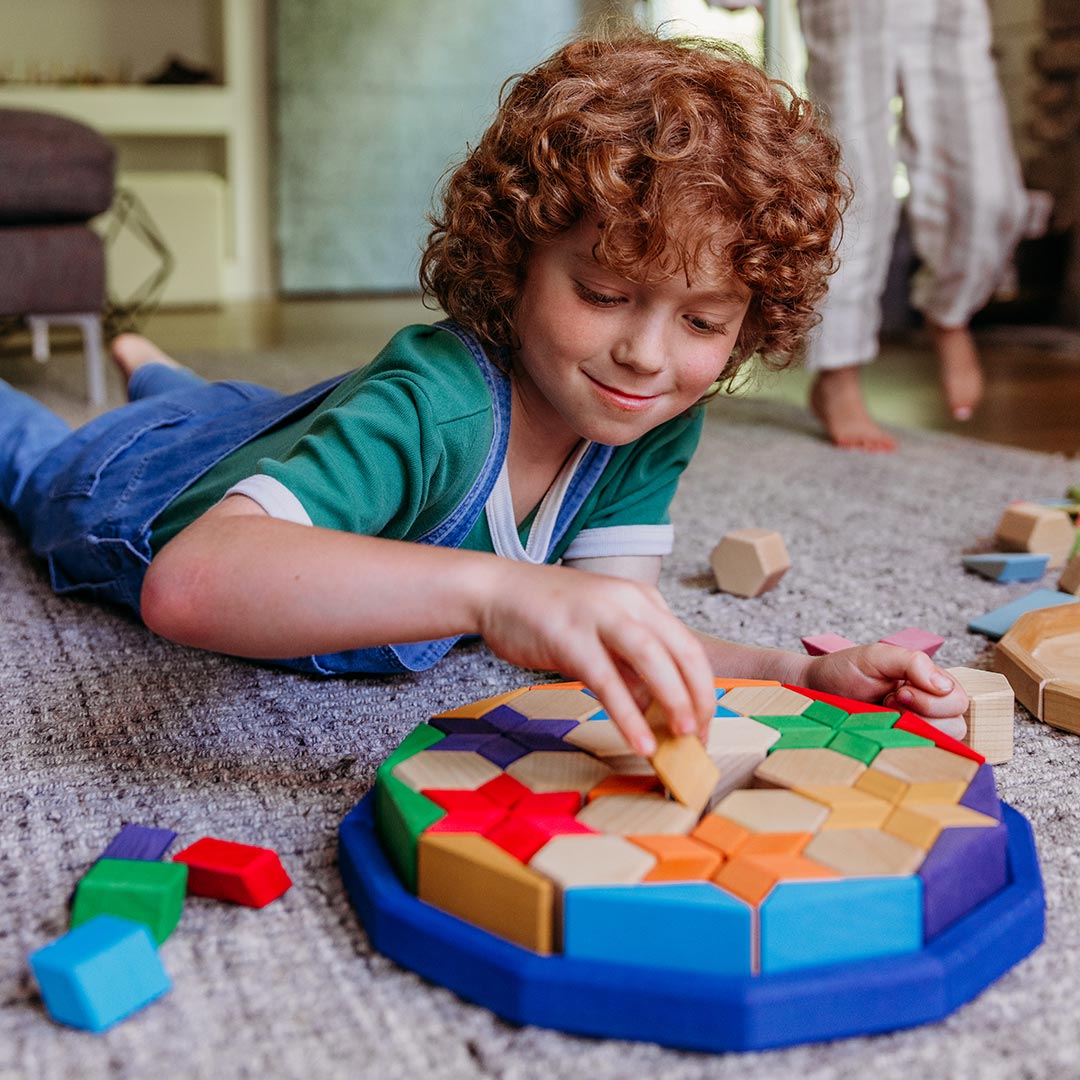 A young child with curly red hair lies on a carpeted floor, arranging colorful geometric wooden blocks on a blue circular tray. The child wears a green shirt and denim overalls in a home setting, with another person's legs visible in the background.