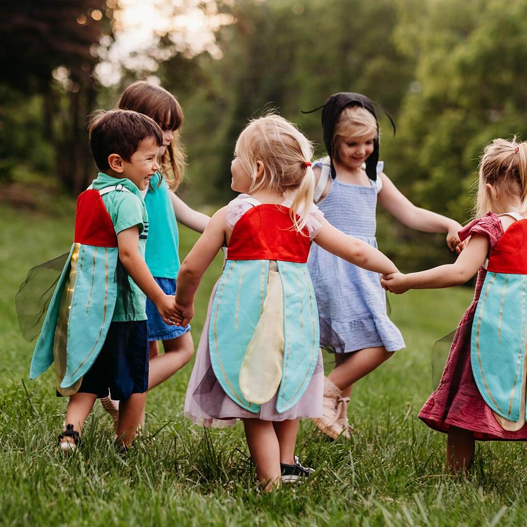 Five children outdoors in a grassy park, holding hands and wearing matching costumes with red tops and large light blue wings with gold trim.
