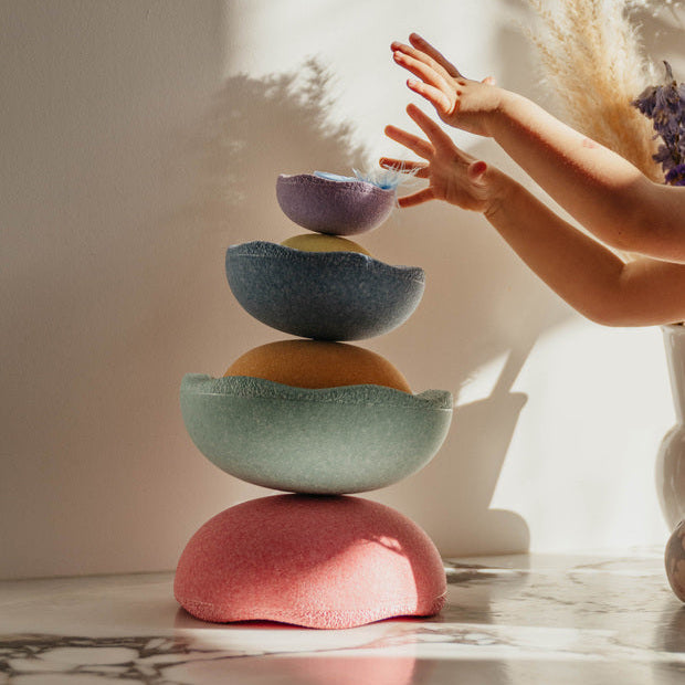 Child's hands reaching toward a tall, balanced stack of pastel-colored Stapelstein® Inside elements in soft morning light, with dried flowers casting shadows in the background.