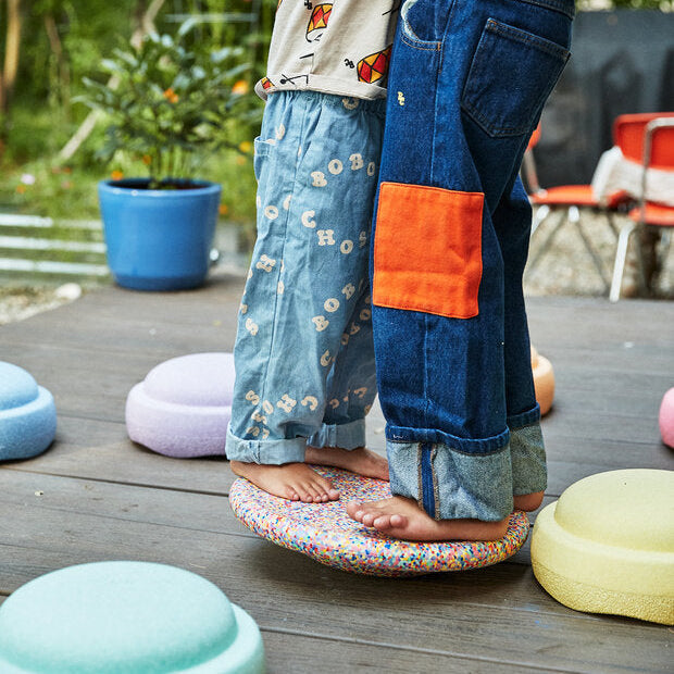 Two children standing barefoot on a Stapelstein® Super Confetti Balance Board outdoors, surrounded by pastel-colored stepping stones on a wooden deck, with plants and play equipment in the background.