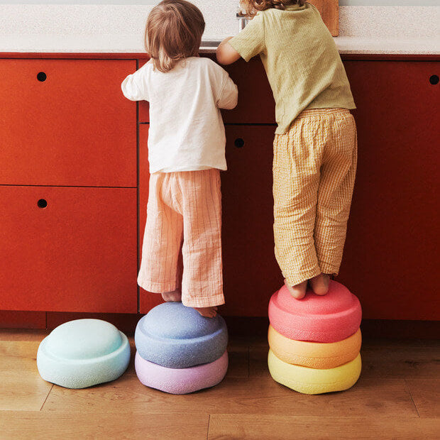 Two young children standing on pastel-colored Stapelstein® stacking elements in front of a red cabinet, using the stones as stools to reach a countertop in a modern indoor setting.