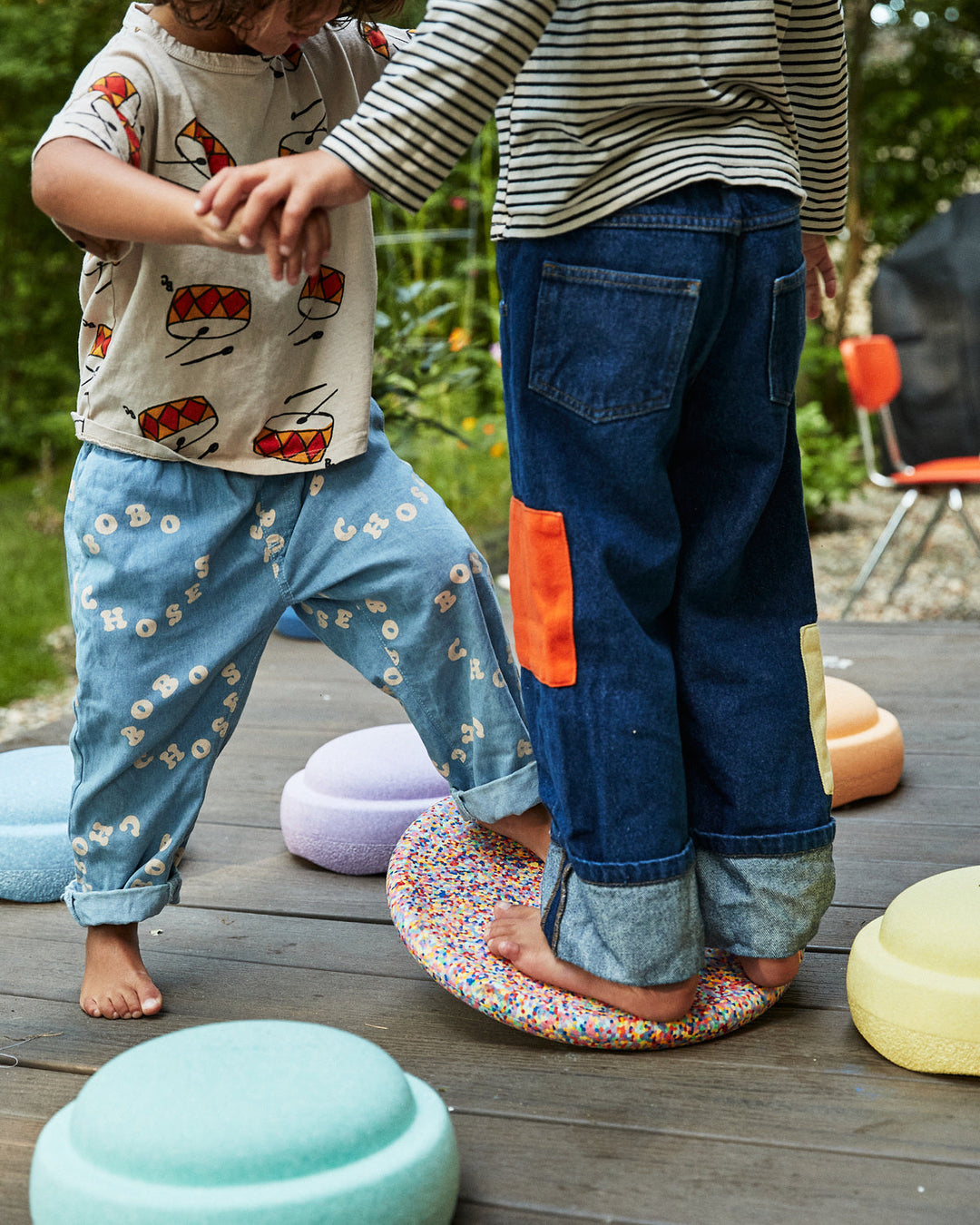 Two children playing outdoors with Stapelstein® elements—one standing barefoot on a Super Confetti stepping stone while holding hands with another child. Pastel-colored stepping stones are scattered on a wooden deck in the background.