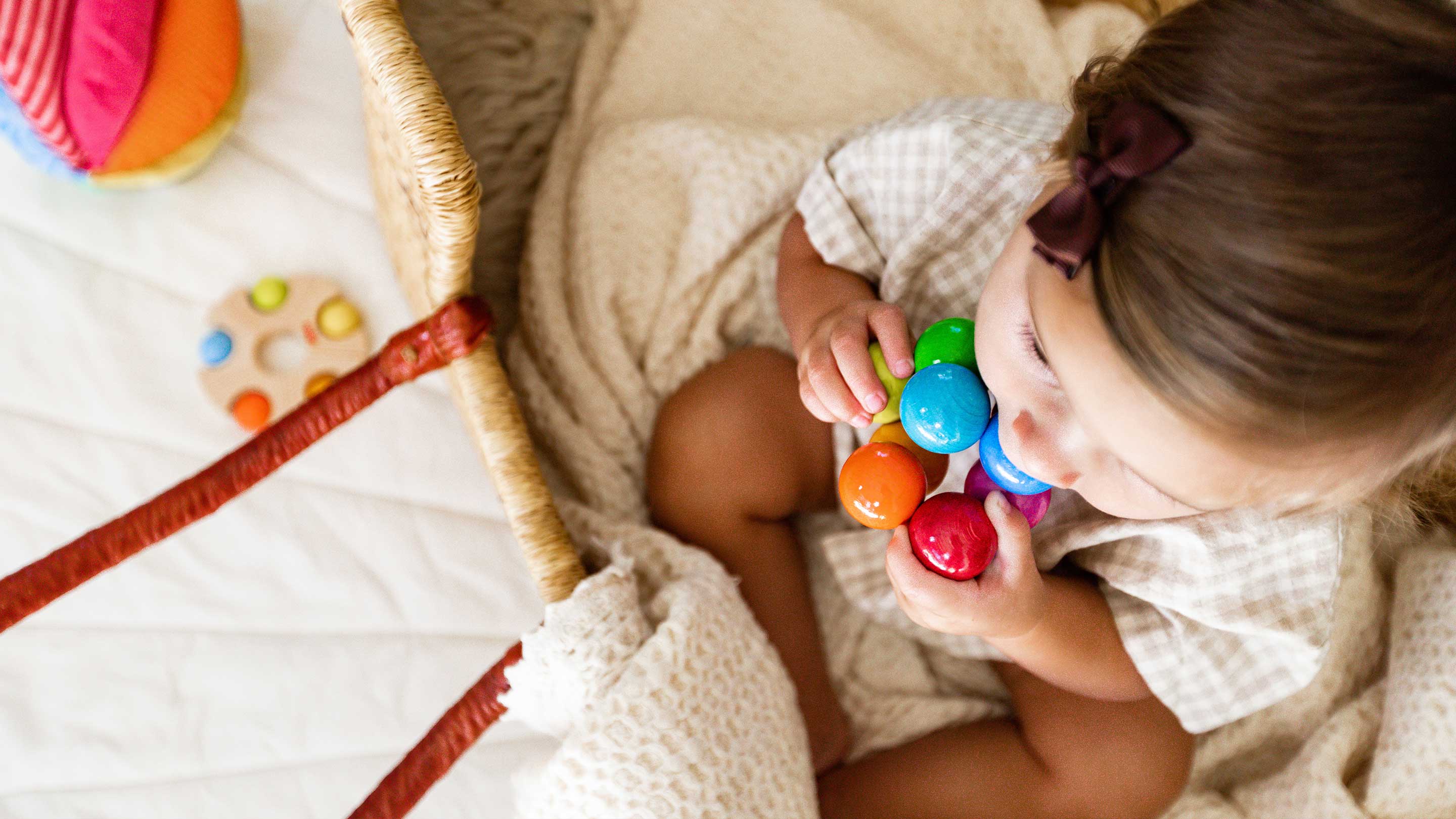A baby wearing a checkered shirt and a dark bow headband sits in a woven bassinet, holding a colorful baby teething toy.