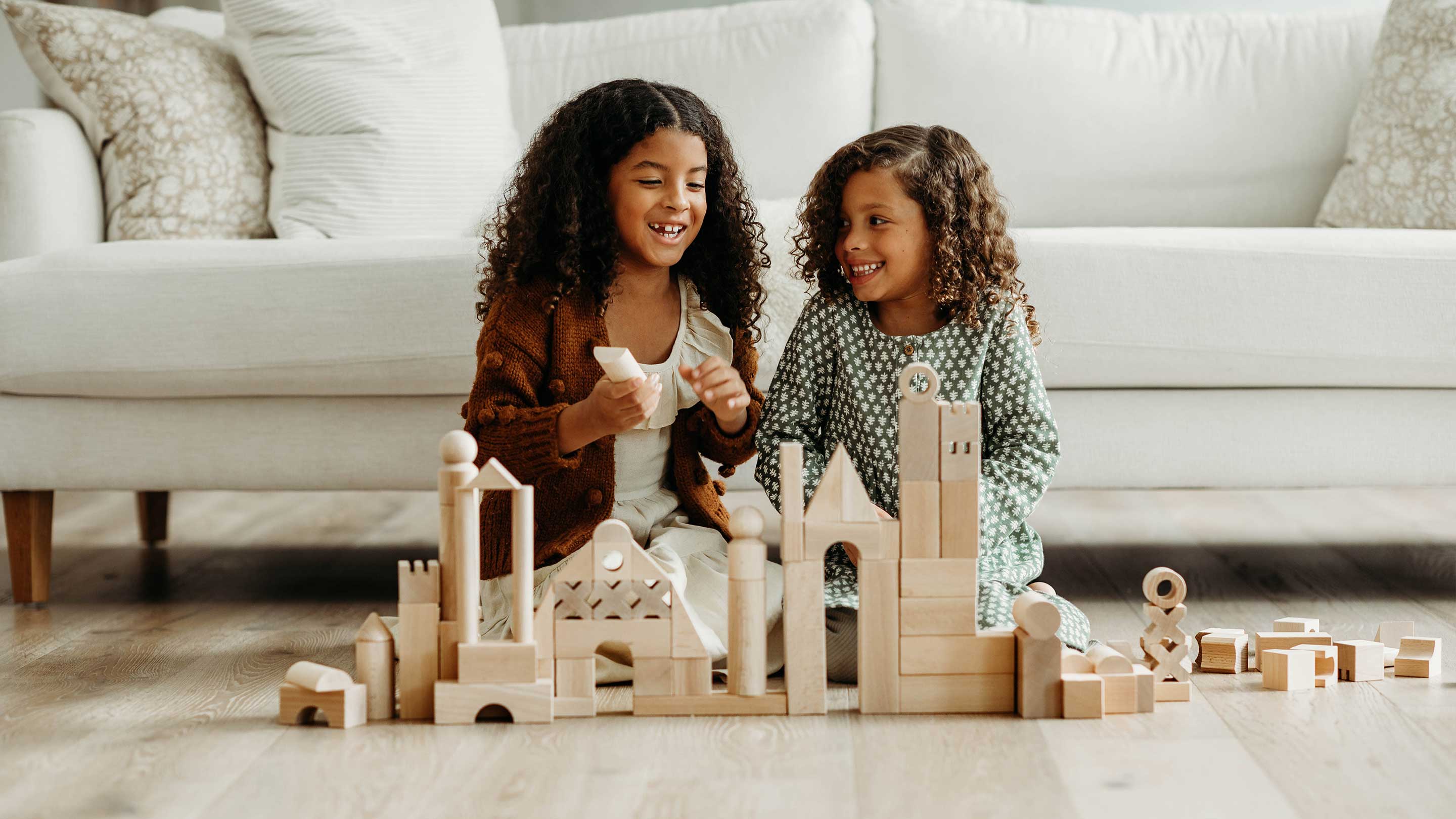 Two young girls smiling and playing with large wooden building blocks on a light wood floor in front of a white sofa.