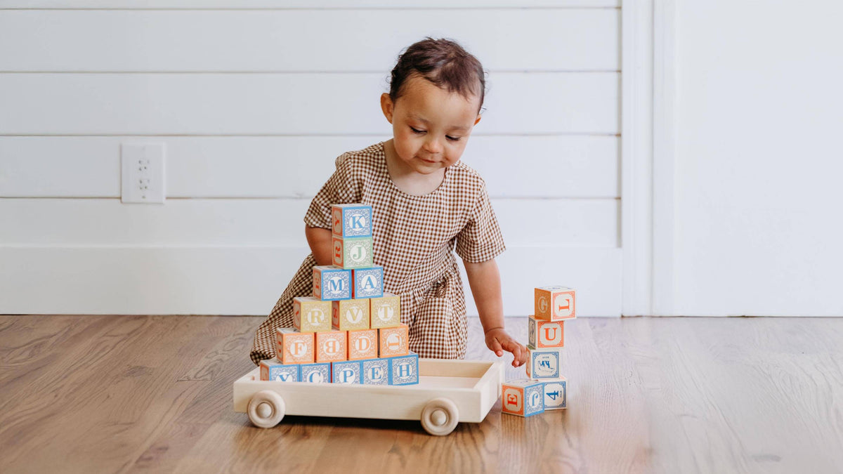 A toddler girl in a brown and beige checkered dress plays with a wooden toy cart loaded with colorful alphabet blocks on a light wood floor against a white paneled wall.