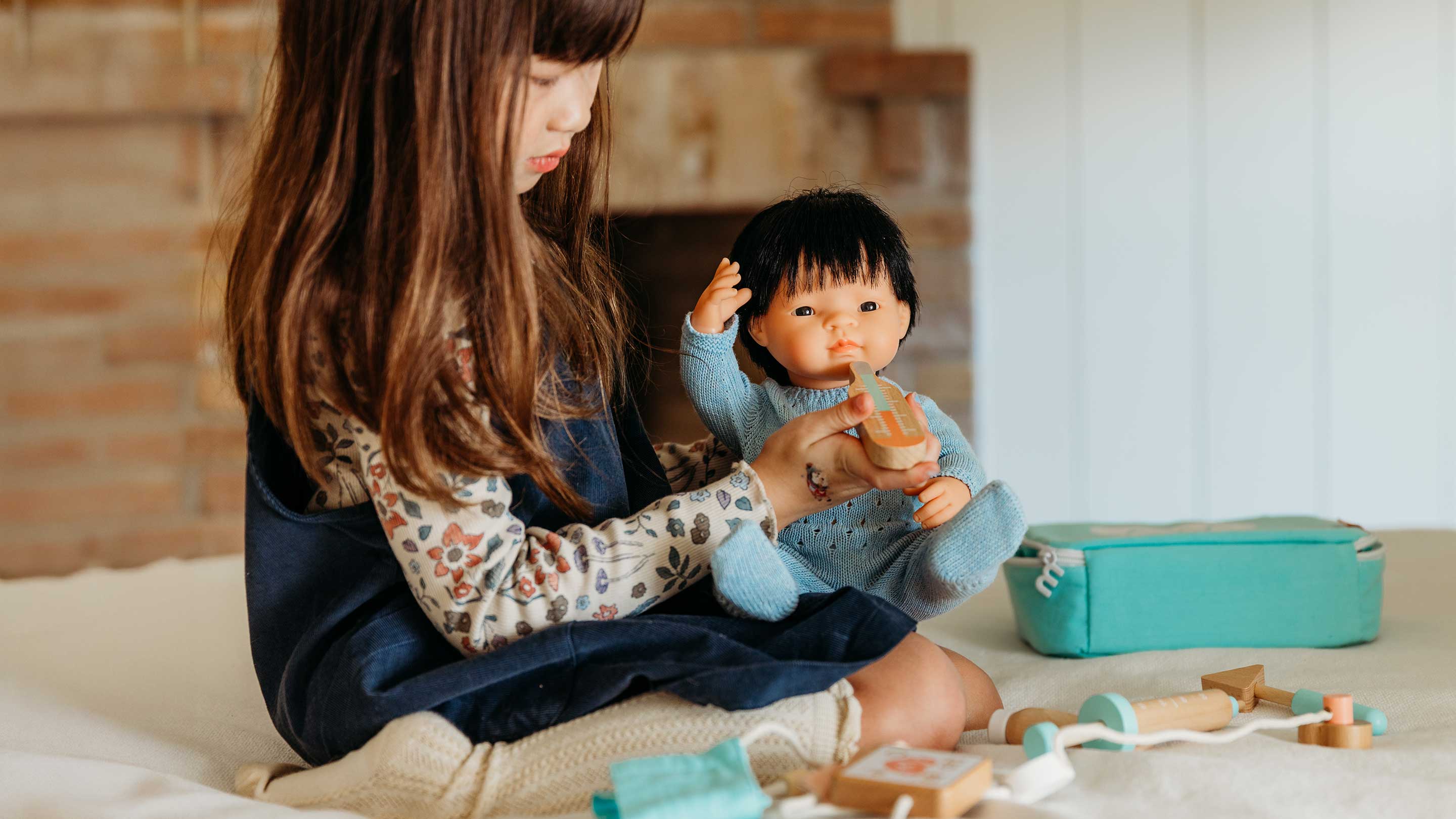 A young girl with long brown hair sits on a cream sofa in a living room, holding a realistic baby doll with black hair and a pacifier. A teal fabric storage bag and wooden toys are on the sofa next to her.
