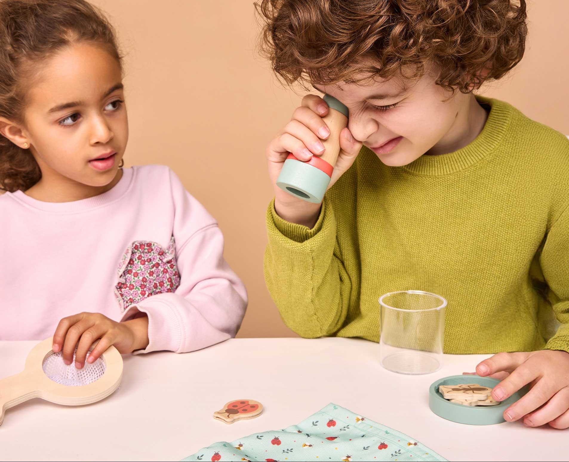 Two children sitting at a table playing with wooden toys. The child on the right wears a green sweater and holds a toy to their eye, while the child on the left wears a pink sweatshirt and looks on.