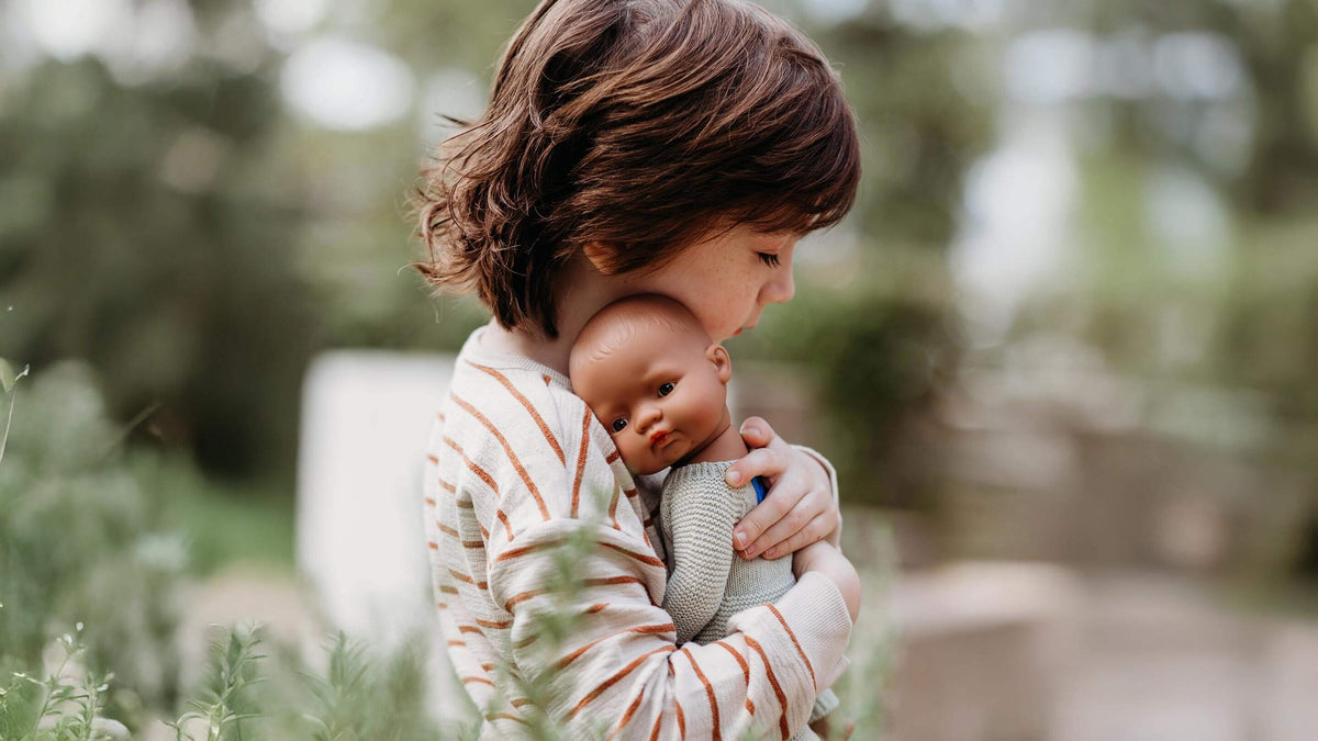 A young child with short brown hair holds a realistic baby doll dressed in a gray outfit, set against a soft-focus outdoor garden background.