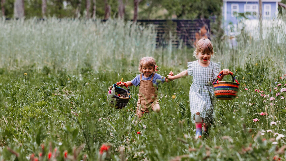Two young children running through a grassy field holding colorful woven baskets, with trees and a house blurred in the background.