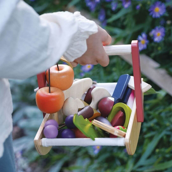 Child's hand holding a toy wooden cart with colorful fruits and vegetables against a blurred natural background.
