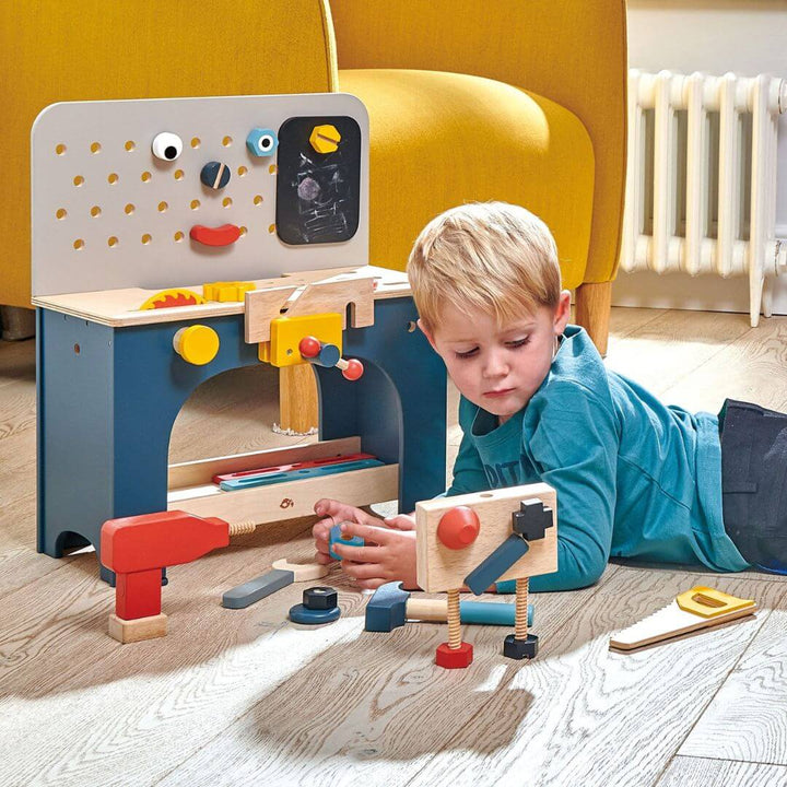 Child playing with a wooden toy workbench in a room with a yellow chair. Tender Leaf Toys