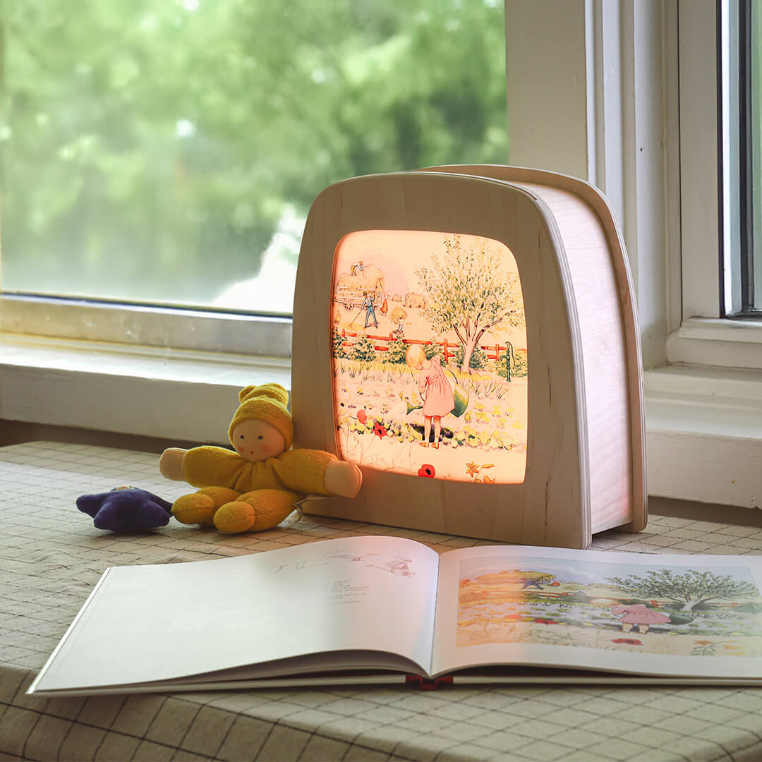 Children's book with a light-up Elsa Beskow Around the Year transparency for Toverlux lamp next to a plush toy on a windowsill.