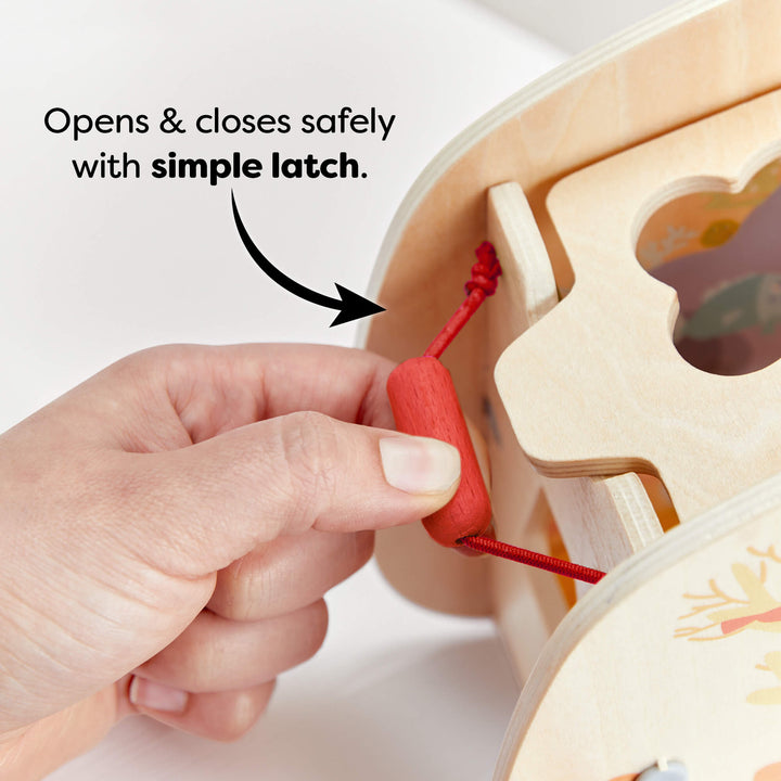 Close-up of a hand opening a simple red latch on a wooden toy shape sorter. Text reads "Opens & closes safely with simple latch."