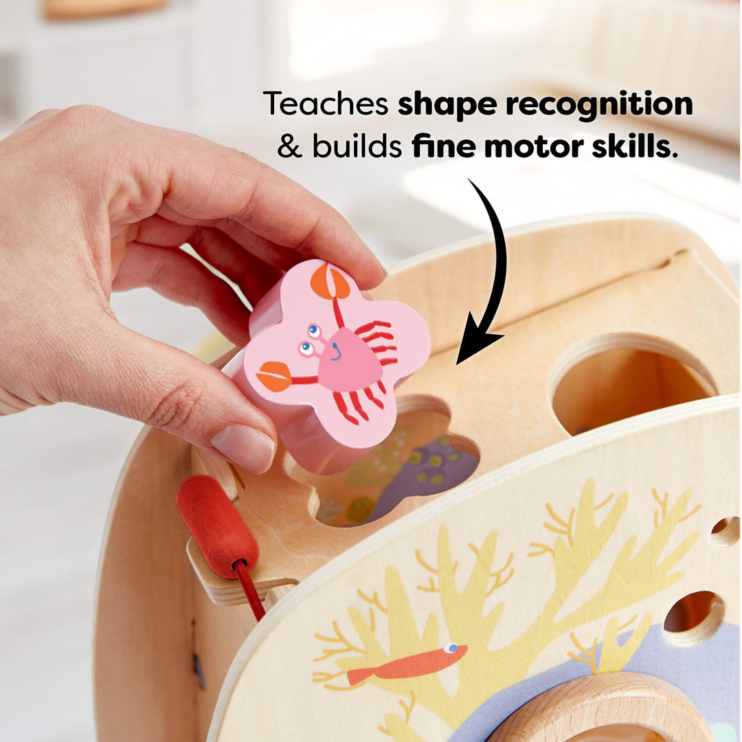 A hand placing a pink crab-shaped block into a matching slot on a wooden toy. Text reads "Teaches shape recognition & builds fine motor skills."