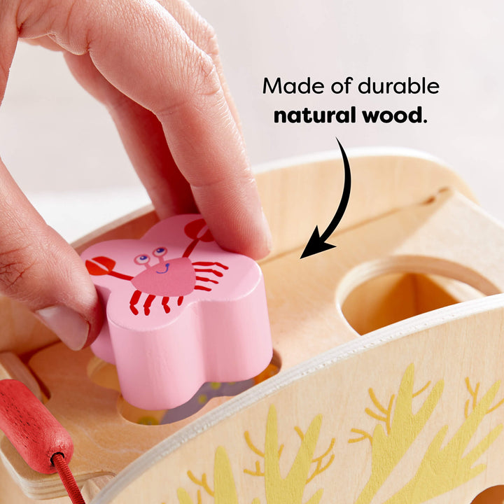Child inserting a pink crab-shaped wooden block into a matching slot on a wooden toy. Text reads "Made of durable natural wood."