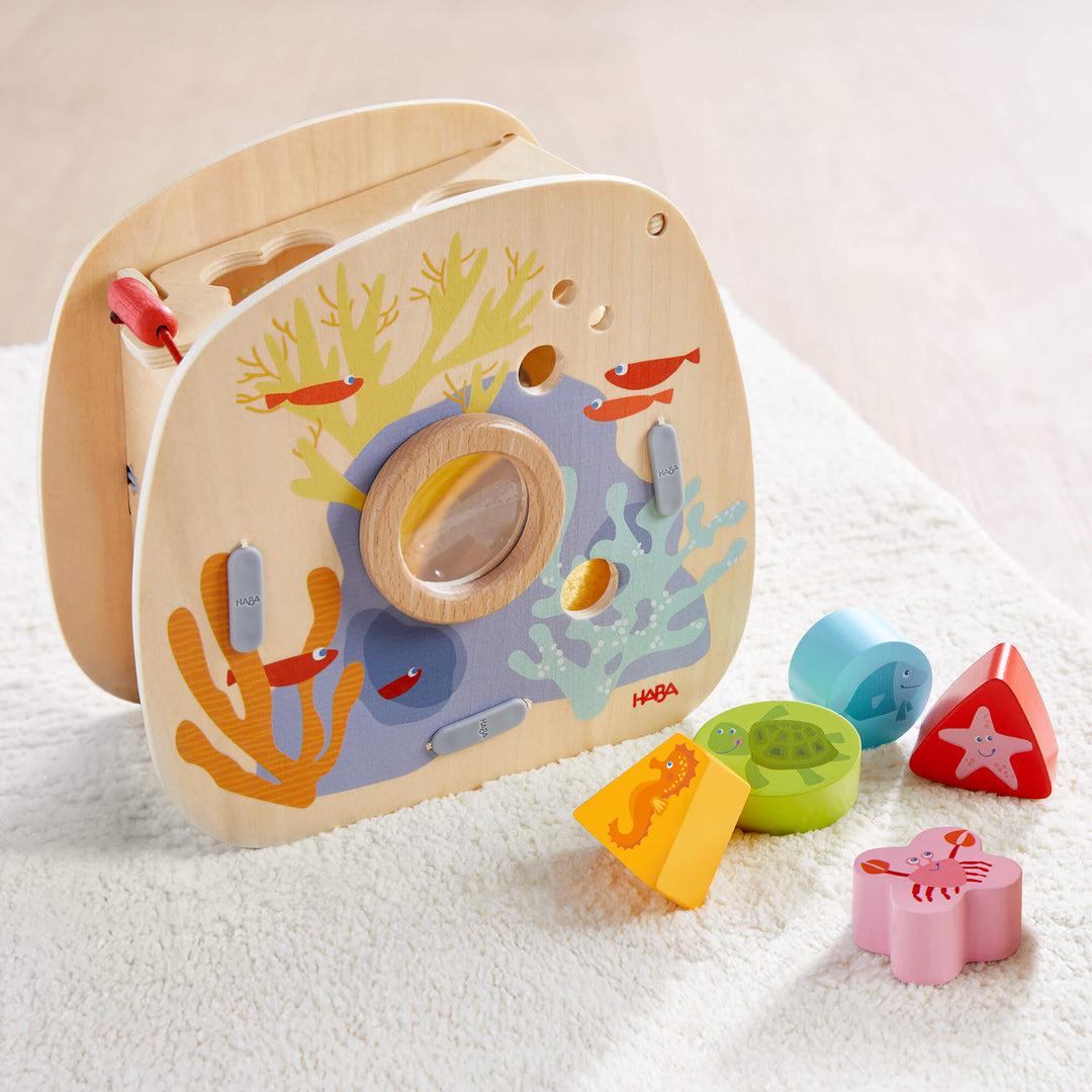 Wooden toy shape sorter with underwater theme, featuring fish and corals, with various shaped blocks in front sitting on a white carpet